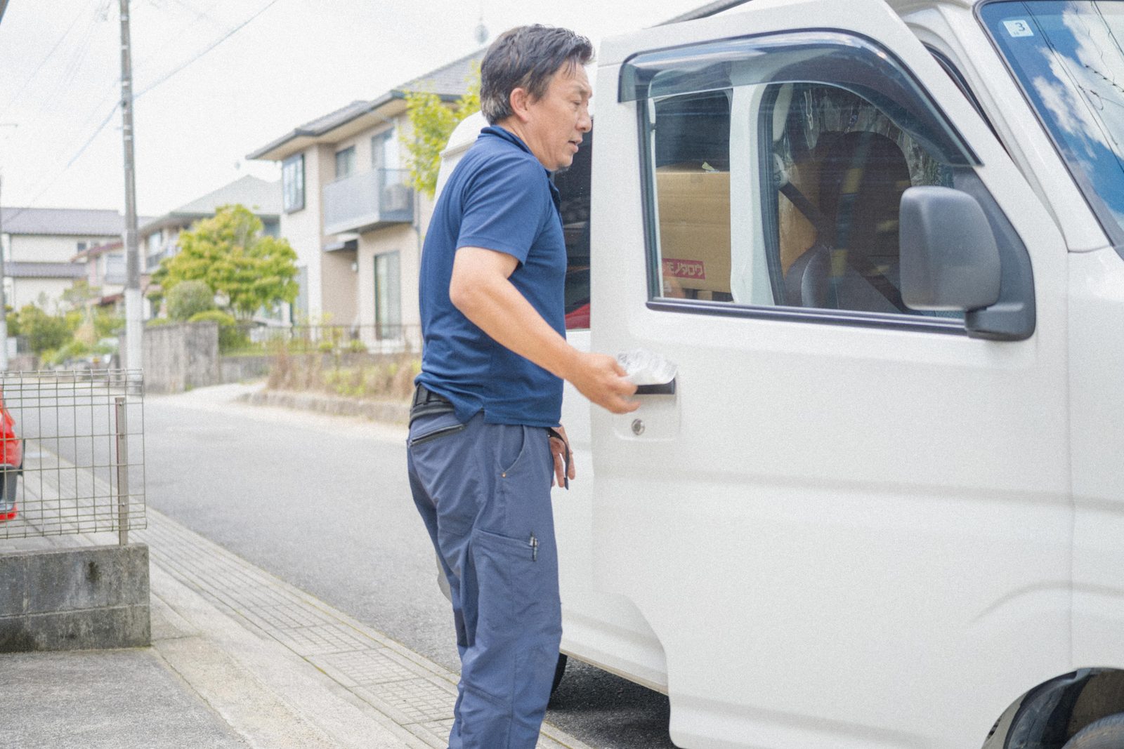 住宅街で白い軽貨物車の運転席ドアの横に立つ I さん（青いポロシャツ）